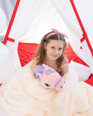 Kıvırcık saçlı, pelüş bir oyuncakla kendini sıcak terli bir battaniyeye sarmış bir kız çocuğu. çocuk odasında rahatla. Yatmadan önce.