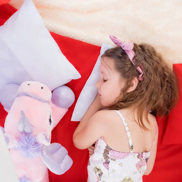 Evde. Tam teşekküllü çocuk uykusu. Uyku maskesi ve pijamalı bir kız kırmızı ve beyaz yastıklarda uyur ve yakınlarda bir tek boynuzlu at peluş oyuncağı yatar..