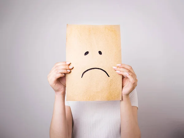 young woman hides her face behind a sheet of paper with a sad smiley face drawn on a gray background,