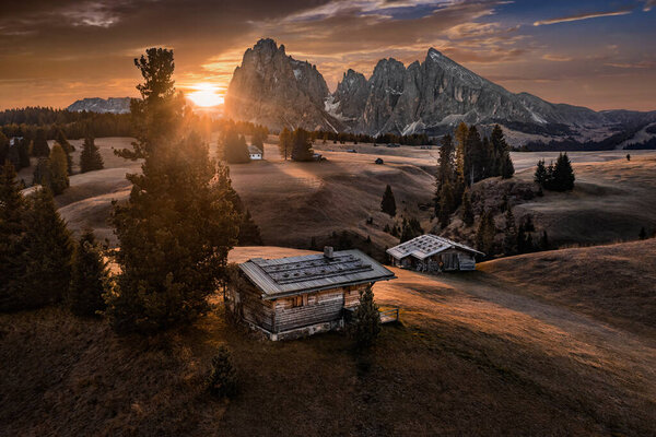 Alpe di Siusi, Italy - Aerial view of wooden cottage at Seiser Alm, a Dolomite plateau in South Tyrol in the Dolomites mountain range at autumn sunrise with colorful sky and the peaks of Kesselkogel