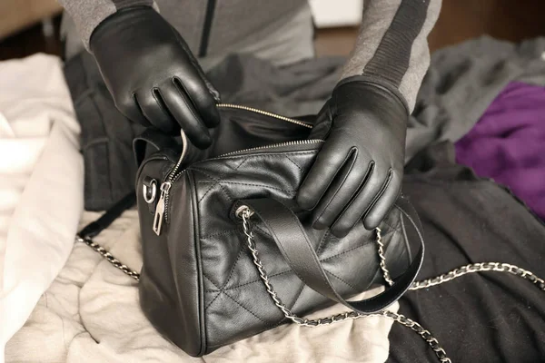 Robber in black outfit and gloves see on opened stolen women bag. A thief evaluates the value of stolen items from a womans handbag in kitchen interior