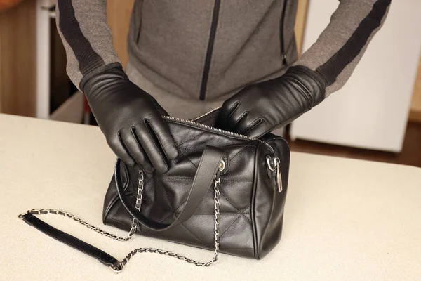 黒の服と手袋で強盗盗まれた女性のバッグを開きます 泥棒は台所のインテリアの女性のハンドバッグから盗品の価値を評価します — ストック写真