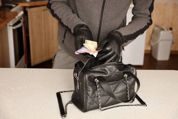 Robber in black outfit and gloves see in opened stolen women bag. The thief takes out the ukrainian money from a womans handbag in kitchen interior