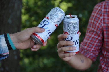 SUMY, UKRAINE - AUGUST 01, 2021: Young man raise Budweiser Bud beer can with male friend on blurred river with kayak and trees. Budweiser is one of the most popular beer brands in the United States