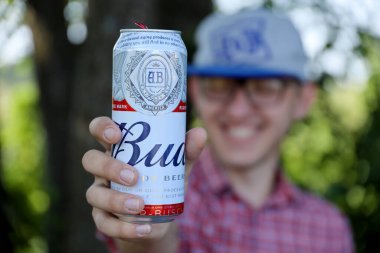 SUMY, UKRAINE - 1 Ağustos 2021: genç adam Budweiser Bud bira kutusunu kaldırdı ve bulanık nehir ve ağaçların arkasında BUD logosu gösterdi. Budweiser, Amerika Birleşik Devletleri 'nin en popüler bira markalarından biridir.
