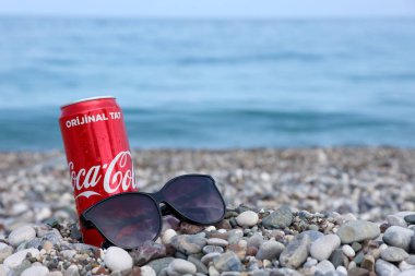 ANTALYA, TURKEY - 18 Mayıs 2021: Orijinal Coca Cola kırmızı teneke kutu deniz kıyısına yakın küçük yuvarlak çakıl taşları üzerinde yatar. Türk sahilinde Coca-Cola kutusu ve güneş gözlüğü