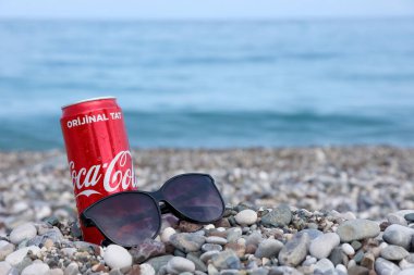 ANTALYA, TURKEY - 18 Mayıs 2021: Orijinal Coca Cola kırmızı teneke kutu deniz kıyısına yakın küçük yuvarlak çakıl taşları üzerinde yatar. Türk sahilinde Coca-Cola kutusu ve güneş gözlüğü