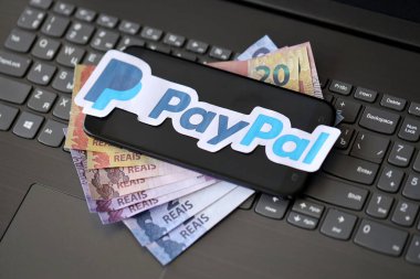 TERNOPIL, UKRAINE - SEPTEMBER 6, 2022 Paypal paper logotype lies on black laptop keyboard with brazilian reals bills. Payoneer is American financial services company provides online money transfer