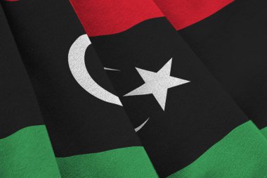 Stüdyo ışıklarının altında büyük kıvrımlı Libya bayrağı sallanıyor. Resmi semboller ve renkler kumaş pankartıyla