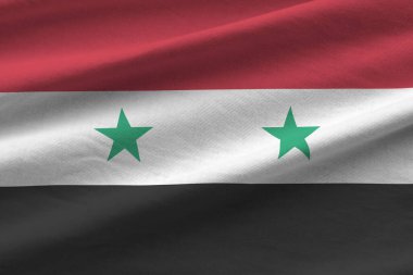 Stüdyo ışıklarının altında büyük kıvrımlı Suriye bayrağı sallanıyor. Resmi semboller ve renkler kumaş pankartıyla