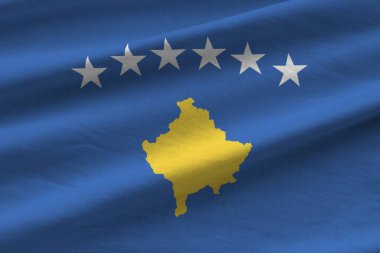 Stüdyo ışıklarının altında kapalı kapılar ardında dalgalanan büyük kıvrımlı Kosova bayrağı. Resmi semboller ve renkler kumaş pankartıyla