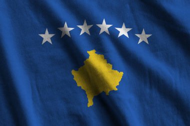 Stüdyo ışıklarının altında kapalı kapılar ardında dalgalanan büyük kıvrımlı Kosova bayrağı. Resmi semboller ve renkler kumaş pankartıyla