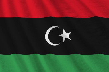 Stüdyo ışıklarının altında büyük kıvrımlı Libya bayrağı sallanıyor. Resmi semboller ve renkler kumaş pankartıyla