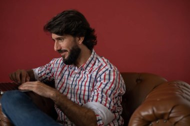 Genç bir iş adamı, arka planda kırmızı bir duvar olan kahverengi bir koltukta dizüstü bilgisayarla çalışıyor. Çalışmak için gri defter. Merkez ofis konsepti. Yüksek kalite fotoğraf