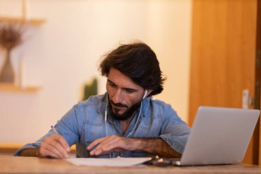 Evde dizüstü bilgisayar ve kağıtlarla çalışan genç bir iş adamı. Çalışanlar için gri defter ve beyaz kulaklıklar. Merkez ofis konsepti. Yüksek kalite fotoğraf