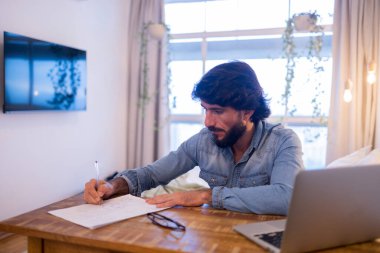 Genç bir iş adamı evinde çalışıyor. Dizüstü bilgisayarı ve masa başında kağıtları var. Çalışmak için gri defter. Merkez ofis konsepti. Yüksek kalite fotoğraf