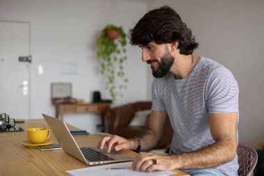 Genç bir iş adamı mutfakta dizüstü bilgisayarla ve mutfak masasında kağıtlarla çalışıyor. Çalışmak için gri defter. Merkez ofis konsepti. Yüksek kalite fotoğraf