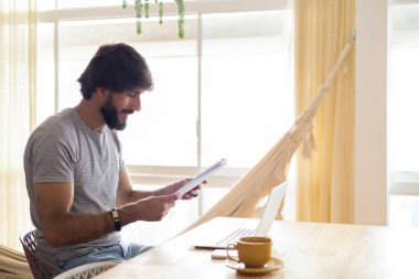 Genç bir iş adamı mutfakta dizüstü bilgisayarla ve mutfak masasında kağıtlarla çalışıyor. Çalışmak için gri defter. Merkez ofis konsepti. Yüksek kalite fotoğraf