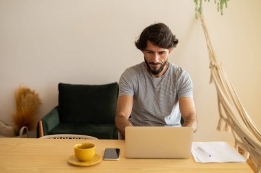 Genç bir iş adamı mutfakta dizüstü bilgisayarla ve mutfak masasında kağıtlarla çalışıyor. Çalışmak için gri defter. Merkez ofis konsepti. Yüksek kalite fotoğraf