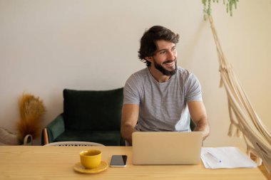Genç bir iş adamı mutfakta dizüstü bilgisayarla ve mutfak masasında kağıtlarla çalışıyor. Çalışmak için gri defter. Merkez ofis konsepti. Yüksek kalite fotoğraf