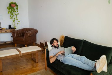 Oturma odasındaki kanepede dizüstü bilgisayarla çalışan genç bir iş adamı. Yerde yatan adam. Çalışmak için gri defter. Yeşil koltuk. Merkez ofis konsepti. Yüksek kalite fotoğraf