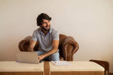 Genç bir iş adamı, arka planda beyaz bir duvar olan kahverengi bir koltukta dizüstü bilgisayarla çalışıyor. Çalışmak için gri defter. Merkez ofis konsepti. Yüksek kalite fotoğraf
