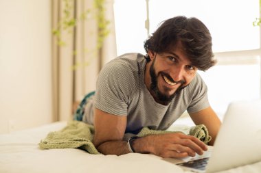 Yatağında dizüstü bilgisayarla yatak odasından çalışan genç bir adam. Yerde yatan adam. Yerde yatan adam. Çalışmak için gri defter. Rahat ve rahat. Merkez ofis konsepti. Yüksek kalite fotoğraf