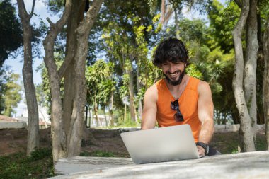 Güzel, güneşli bir günde kucağında dizüstü bilgisayarla parkta genç bir adam. Çalışmak ve boş vakit geçirmek için gri defter. Merkez ofis konsepti. Yeşil ve doğa geçmişi. Yüksek kalite fotoğraf
