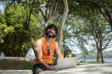 Güzel, güneşli bir günde kucağında dizüstü bilgisayarla parkta genç bir adam. Çalışmak ve boş vakit geçirmek için gri defter. Merkez ofis konsepti. Yeşil ve doğa geçmişi. Yüksek kalite fotoğraf