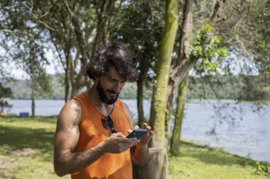 Güzel güneşli bir günde, cep telefonuyla parkta genç bir adam. Çalışmak ve rahatlamak için cep telefonu. Merkez ofis konsepti. Yeşil ve doğa geçmişi. Yüksek kalite fotoğraf