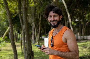 Güzel güneşli bir günde, cep telefonuyla parkta genç bir adam. Çalışmak ve rahatlamak için cep telefonu. Merkez ofis konsepti. Yeşil ve doğa geçmişi. Yüksek kalite fotoğraf