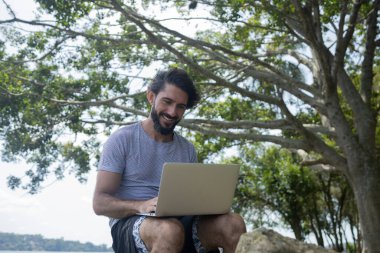 Güzel, güneşli bir günde kucağında dizüstü bilgisayarla parkta genç bir adam. Çalışmak ve boş vakit geçirmek için gri defter. Merkez ofis konsepti. Yeşil ve doğa geçmişi. Yüksek kalite fotoğraf