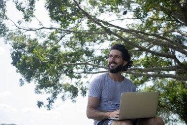 Güzel, güneşli bir günde kucağında dizüstü bilgisayarla parkta genç bir adam. Çalışmak ve boş vakit geçirmek için gri defter. Merkez ofis konsepti. Yeşil ve doğa geçmişi. Yüksek kalite fotoğraf