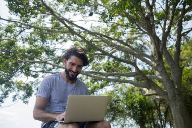 Güzel, güneşli bir günde kucağında dizüstü bilgisayarla parkta genç bir adam. Çalışmak ve boş vakit geçirmek için gri defter. Merkez ofis konsepti. Yeşil ve doğa geçmişi. Yüksek kalite fotoğraf