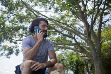Güzel güneşli bir günde, cep telefonuyla parkta genç bir adam. Çalışmak ve rahatlamak için cep telefonu. Merkez ofis konsepti. Yeşil ve doğa geçmişi. Yüksek kalite fotoğraf