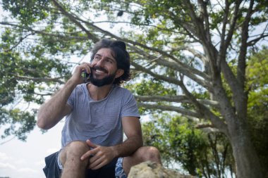 Güzel güneşli bir günde, cep telefonuyla parkta genç bir adam. Çalışmak ve rahatlamak için cep telefonu. Merkez ofis konsepti. Yeşil ve doğa geçmişi. Yüksek kalite fotoğraf