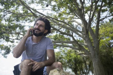 Güzel güneşli bir günde, cep telefonuyla parkta genç bir adam. Çalışmak ve rahatlamak için cep telefonu. Merkez ofis konsepti. Yeşil ve doğa geçmişi. Yüksek kalite fotoğraf