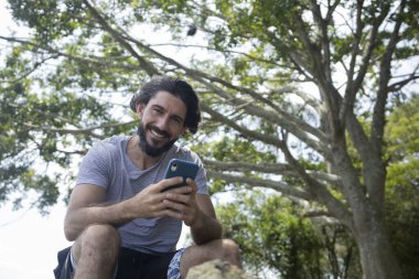 Güzel güneşli bir günde, cep telefonuyla parkta genç bir adam. Çalışmak ve rahatlamak için cep telefonu. Merkez ofis konsepti. Yeşil ve doğa geçmişi. Yüksek kalite fotoğraf