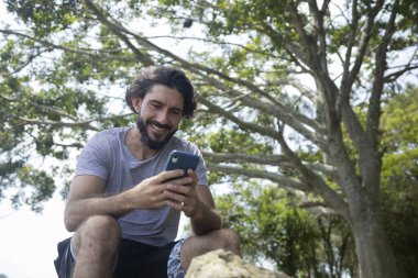Güzel güneşli bir günde, cep telefonuyla parkta genç bir adam. Çalışmak ve rahatlamak için cep telefonu. Merkez ofis konsepti. Yeşil ve doğa geçmişi. Yüksek kalite fotoğraf
