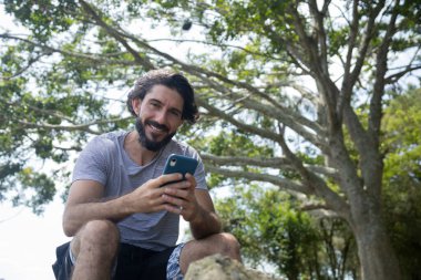 Güzel güneşli bir günde, cep telefonuyla parkta genç bir adam. Çalışmak ve rahatlamak için cep telefonu. Merkez ofis konsepti. Yeşil ve doğa geçmişi. Yüksek kalite fotoğraf