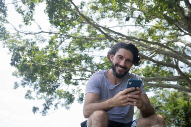 Güzel güneşli bir günde, cep telefonuyla parkta genç bir adam. Çalışmak ve rahatlamak için cep telefonu. Merkez ofis konsepti. Yeşil ve doğa geçmişi. Yüksek kalite fotoğraf