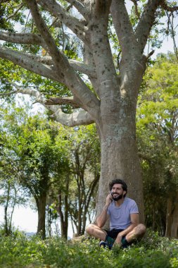 Güzel güneşli bir günde, cep telefonuyla parkta genç bir adam. Çalışmak ve rahatlamak için cep telefonu. Merkez ofis konsepti. Yeşil ve doğa geçmişi. Yüksek kalite fotoğraf