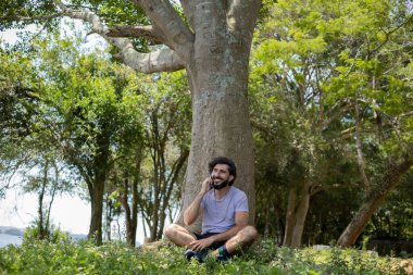 Güzel güneşli bir günde, cep telefonuyla parkta genç bir adam. Çalışmak ve rahatlamak için cep telefonu. Merkez ofis konsepti. Yeşil ve doğa geçmişi. Yüksek kalite fotoğraf