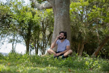 Güzel güneşli bir günde, cep telefonuyla parkta genç bir adam. Çalışmak ve rahatlamak için cep telefonu. Merkez ofis konsepti. Yeşil ve doğa geçmişi. Yüksek kalite fotoğraf