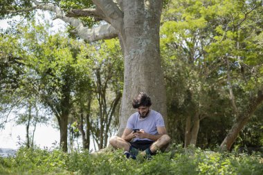 Güzel güneşli bir günde, cep telefonuyla parkta genç bir adam. Çalışmak ve rahatlamak için cep telefonu. Merkez ofis konsepti. Yeşil ve doğa geçmişi. Yüksek kalite fotoğraf