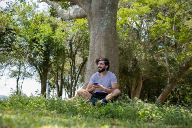 Güzel güneşli bir günde, cep telefonuyla parkta genç bir adam. Çalışmak ve rahatlamak için cep telefonu. Merkez ofis konsepti. Yeşil ve doğa geçmişi. Yüksek kalite fotoğraf