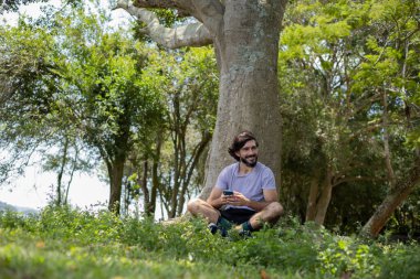 Güzel güneşli bir günde, cep telefonuyla parkta genç bir adam. Çalışmak ve rahatlamak için cep telefonu. Merkez ofis konsepti. Yeşil ve doğa geçmişi. Yüksek kalite fotoğraf