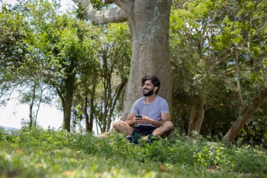 Güzel güneşli bir günde, cep telefonuyla parkta genç bir adam. Çalışmak ve rahatlamak için cep telefonu. Merkez ofis konsepti. Yeşil ve doğa geçmişi. Yüksek kalite fotoğraf