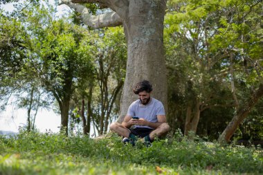 Güzel güneşli bir günde, cep telefonuyla parkta genç bir adam. Çalışmak ve rahatlamak için cep telefonu. Merkez ofis konsepti. Yeşil ve doğa geçmişi. Yüksek kalite fotoğraf