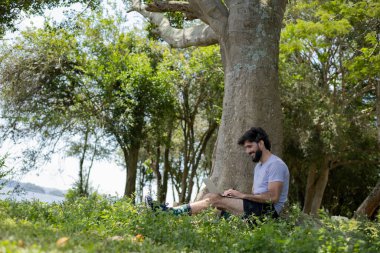 Güzel, güneşli bir günde kucağında dizüstü bilgisayarla parkta genç bir adam. Çalışmak ve boş vakit geçirmek için gri defter. Merkez ofis konsepti. Yeşil ve doğa geçmişi. Yüksek kalite fotoğraf